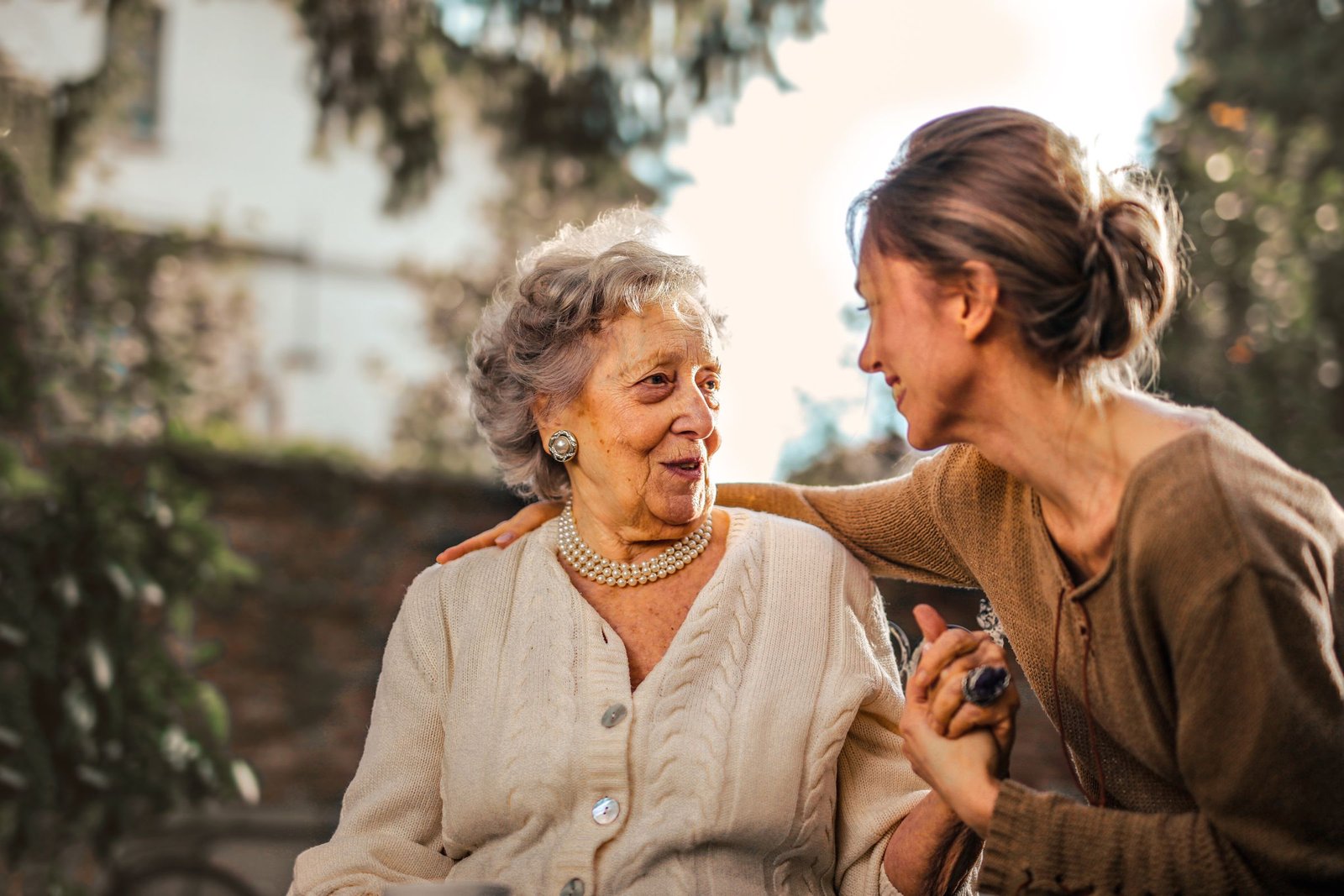 An older woman in a cream cardigan and a younger woman in a brown sweater sit outdoors and hold hands